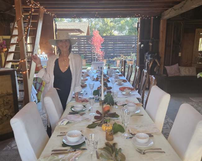 A woman stands next to a long dining table set with dishes and decorations in a rustic, wooden interior space.