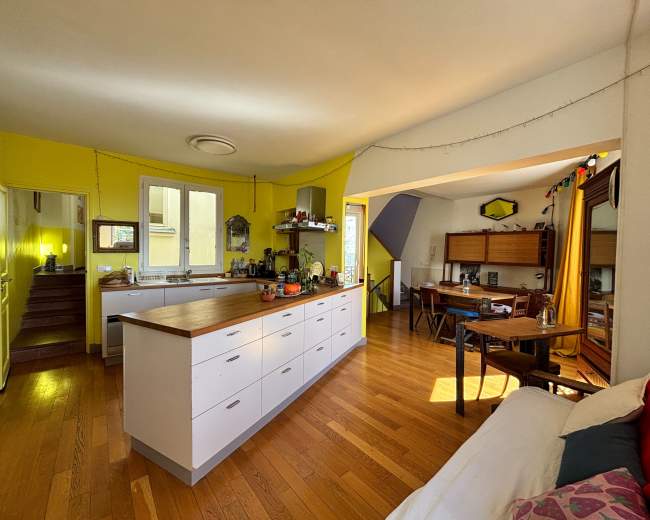The image depicts a brightly lit kitchen and living area with wooden floors, a central island countertop, and a cozy dining space in the background.
