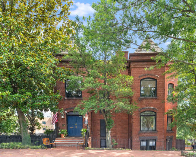 The image shows a two-story brick house with large windows, surrounded by trees and a small garden.
