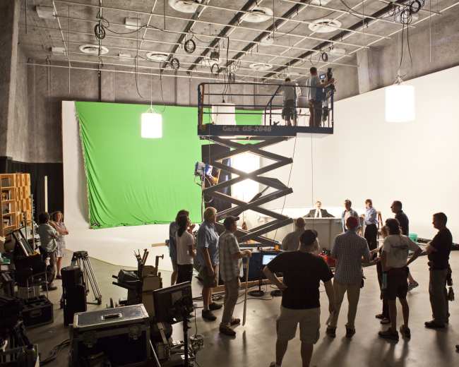 A film crew is set up in a studio, with a scissor lift in the center and a green screen backdrop on one side.