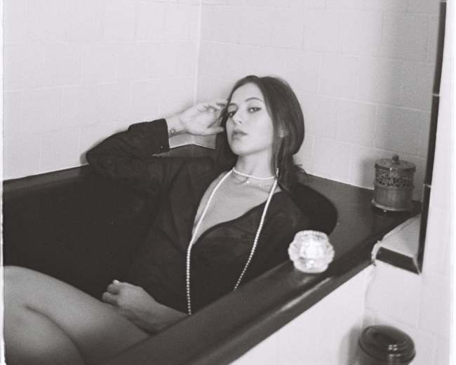 A woman dressed in a black shirt and a pearl necklace is reclining in a vintage bathtub with tiled walls.