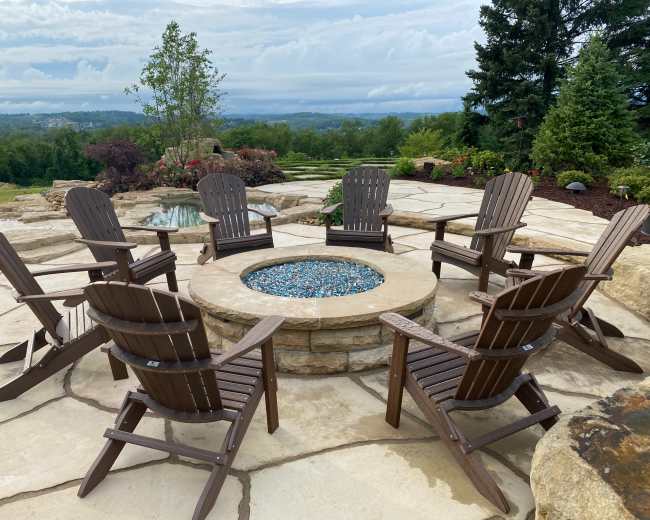 A stone fire pit surrounded by eight wooden chairs sits on a paved patio overlooking a valley and lush greenery.