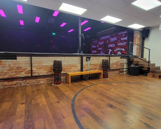 The image shows a spacious room with wooden flooring, a stage area with colorful lighting, and a backdrop featuring graffiti-style art, along with two drum-like instruments and a bench.