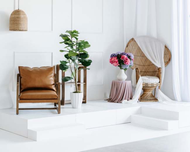 The image features a minimalist indoor setting with a brown leather chair, a rattan chair draped in fabric, a potted plant, and a vase of flowers on a white pedestal, all against a light-colored wall.