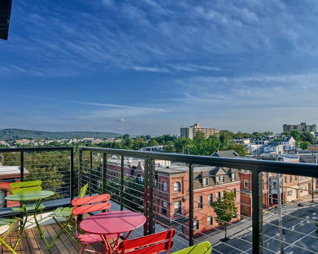 A rooftop balcony features colorful seating and offers a view of a cityscape with buildings and a distant mountain under a clear sky.