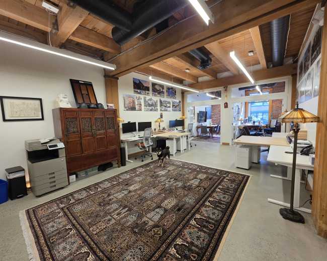 The image shows a spacious office with wooden beams, multiple workstations, a patterned rug, and a dog standing on the carpet.