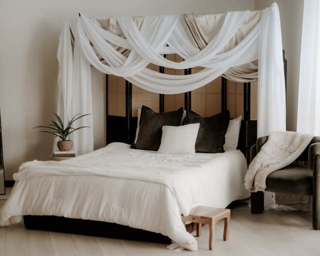 The image depicts a neatly arranged bedroom featuring a large bed with draped white fabric above, flanked by a mirror and a potted plant.