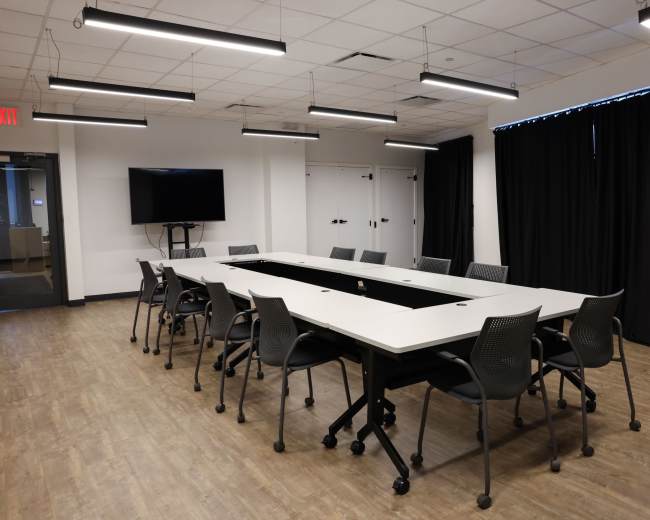 The image shows a modern conference room with a long table surrounded by chairs, featuring a television on the wall and black curtains covering windows.