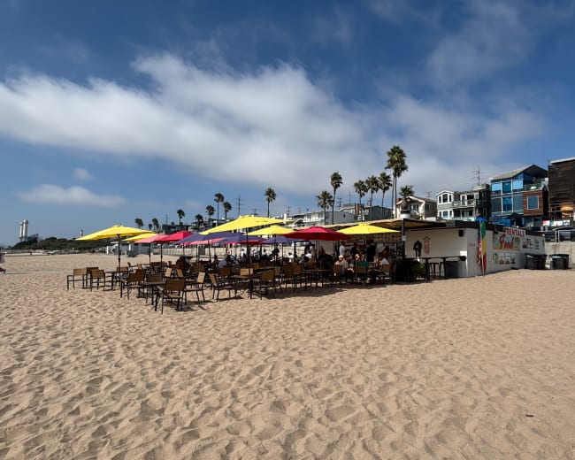 A beachside café with colorful umbrellas provides outdoor seating on sandy shores, surrounded by palm trees and coastal buildings.