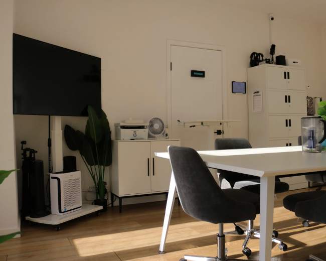 The image shows a modern office space with a white table and gray chairs, a television mounted on the wall, and a storage cabinet.