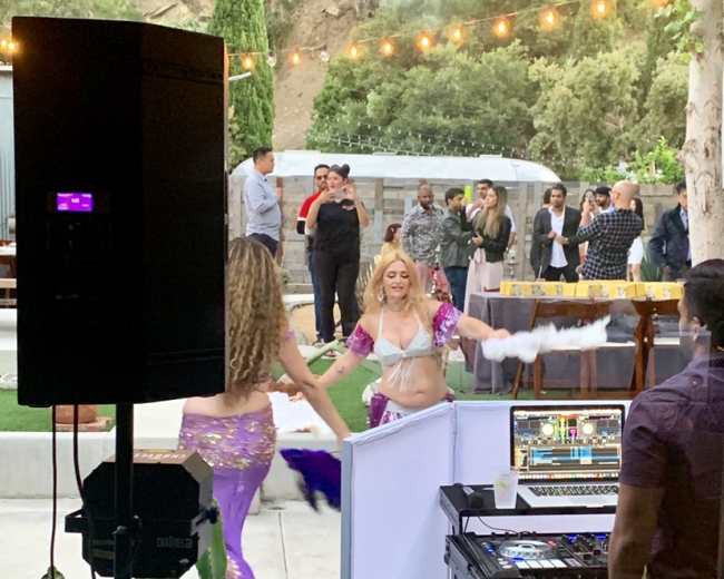 Two dancers in colorful costumes perform in front of an audience at an outdoor event with string lights and greenery in the background.