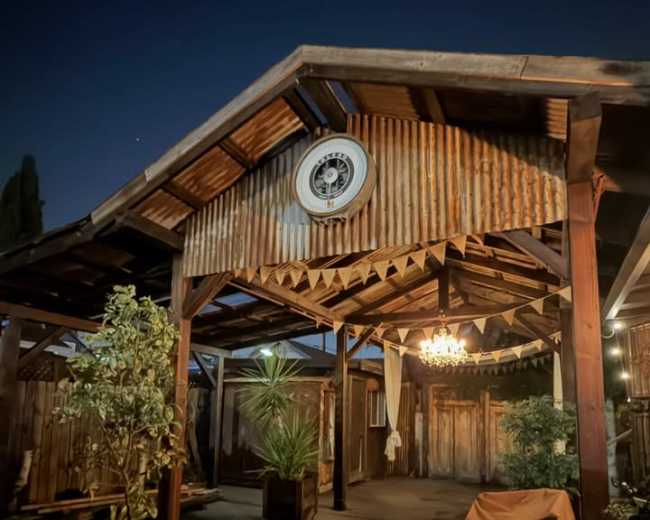 The image shows a rustic outdoor patio with a corrugated metal roof, wooden beams, potted plants, and a chandelier hanging from the ceiling.