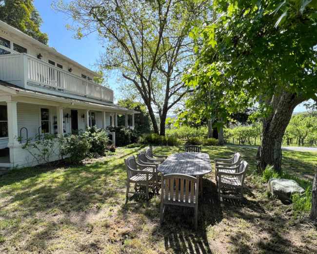A large wooden table with eight chairs is set in a grassy area beside a house with a porch, surrounded by trees and vineyards.