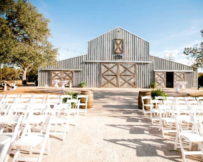 A rustic barn with a gravel path leading to it, surrounded by white folding chairs arranged for an outdoor event.