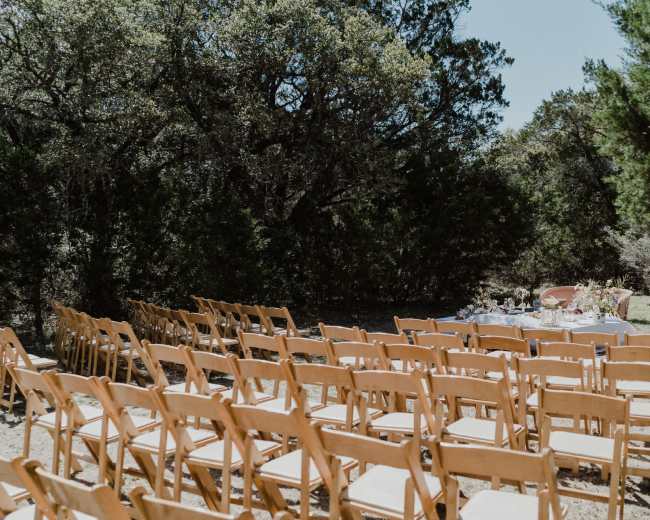 A row of wooden chairs is set up for an outdoor event, facing a table adorned with decor amid a wooded area.