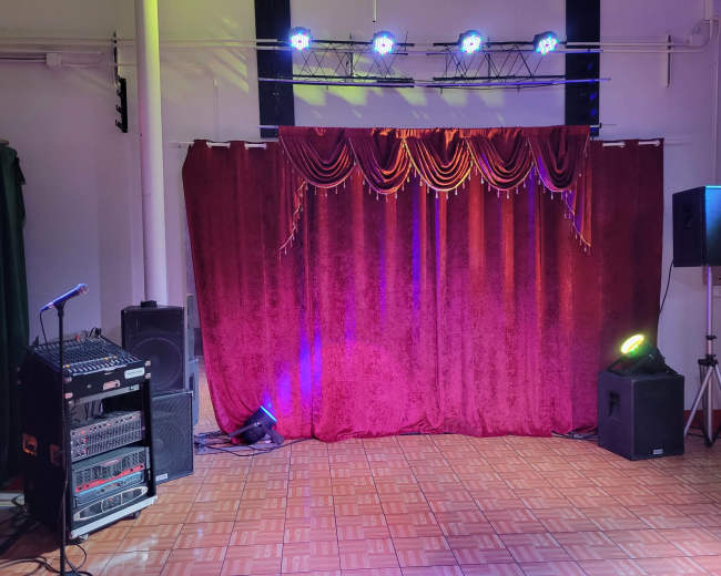 The image shows a stage area with a red curtain draped at the front, flanked by audio equipment and stage lights.