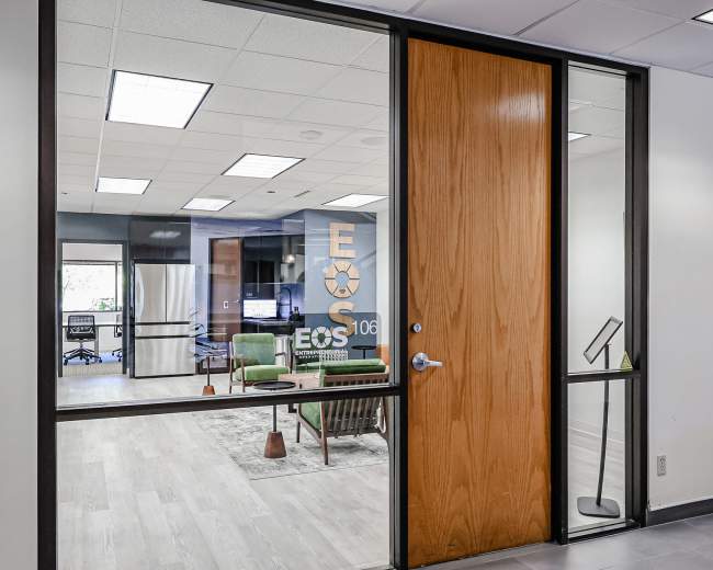 The image shows an office space with a glass door labeled "EOS 106," featuring a modern interior with seating and a kitchenette visible through the window.