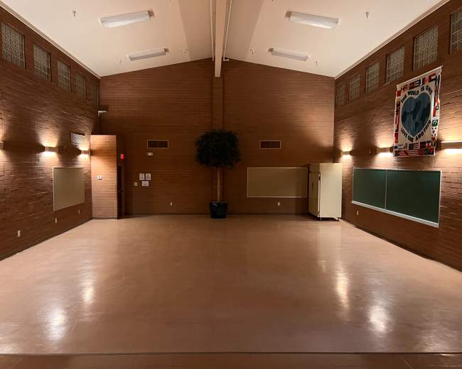 The image shows a spacious, empty room with a polished floor and brown brick walls, featuring a potted plant and two large whiteboards.