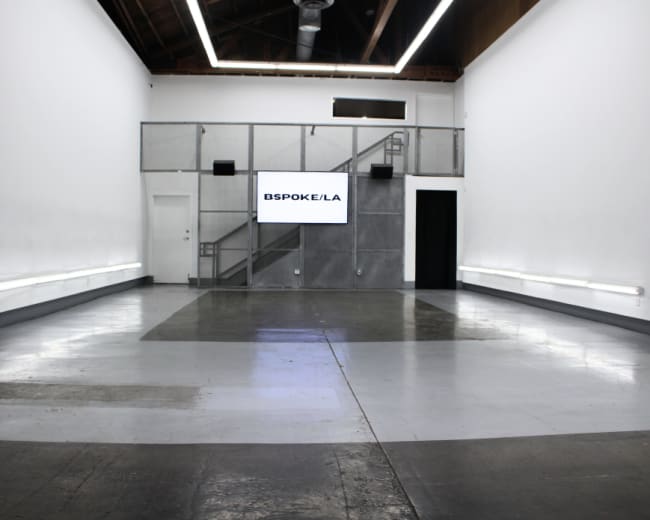 The image shows an empty, well-lit interior space with a large screen displaying "BESPOKE/LA" and a staircase in the background.