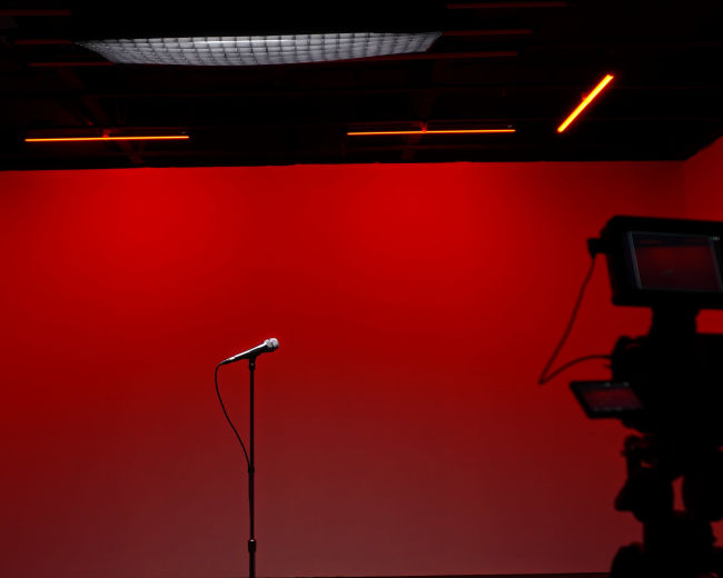 A microphone stands on a black stand in front of a solid red backdrop, with a camera silhouette capturing the scene.