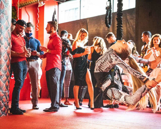 A dancer performs on aerial silks in a lively venue filled with guests mingling and enjoying drinks.