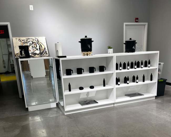 A white shelving unit displays black bottles, with a mirror and two large containers visible in the background of a modern, minimalist room.
