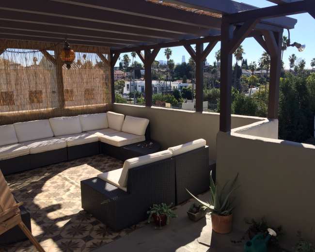 A covered outdoor seating area with a sectional sofa and potted plants overlooks a neighborhood with palm trees in the background.