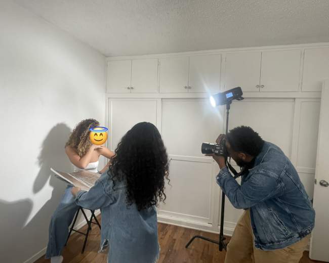 A photographer captures an image of a model sitting on a stool in a well-lit room with white walls and a visible backdrop.