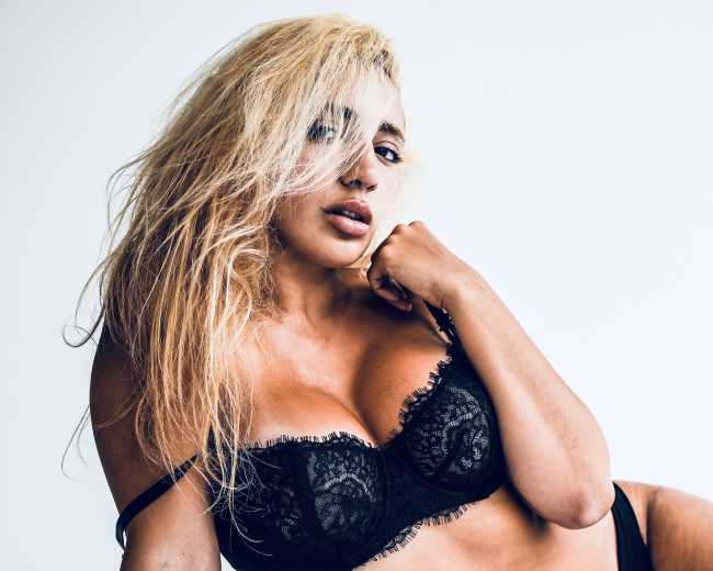 A woman with long, blonde hair poses against a plain white background while wearing a black lace bra.