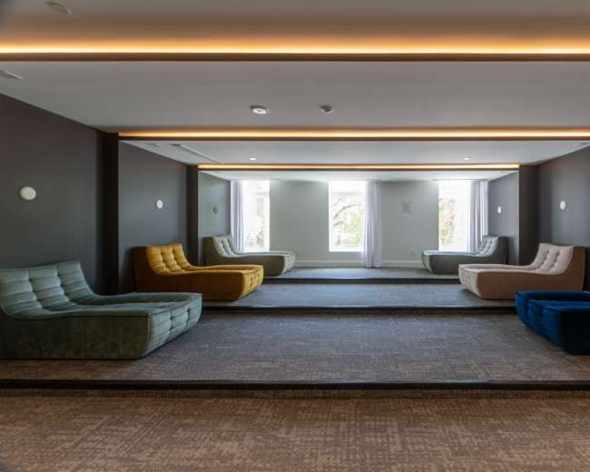 The image shows a modern lounge area featuring several chaise lounges in various colors arranged symmetrically around a central space, with large windows allowing natural light to enter.