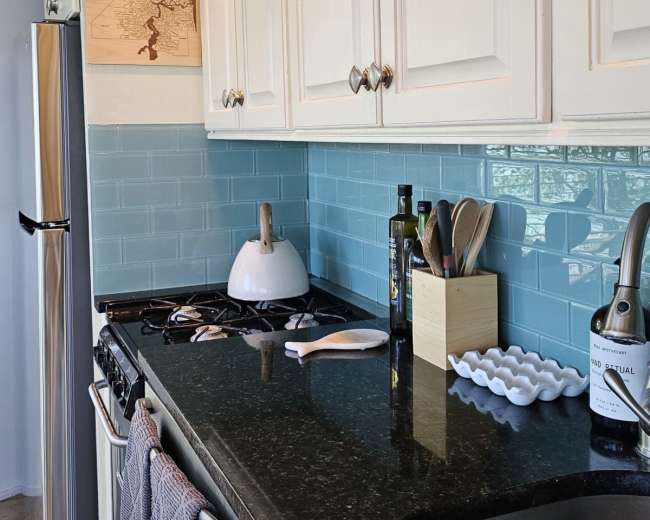 The image shows a modern kitchen with white cabinets, a black countertop, and a light blue backsplash, featuring a stovetop, sink, and various kitchen utensils.