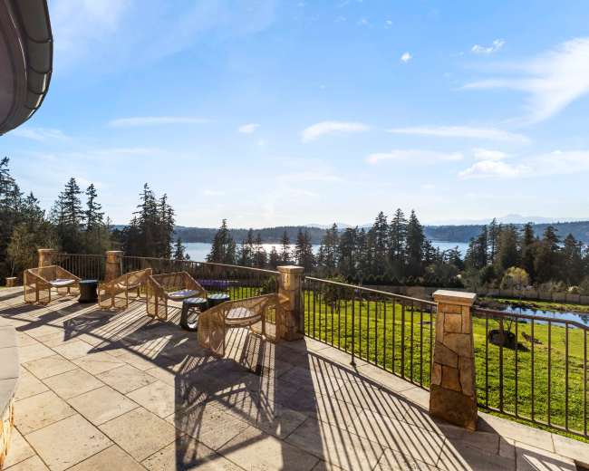 A stone patio with several lounge chairs overlooks a body of water surrounded by trees under a clear blue sky.