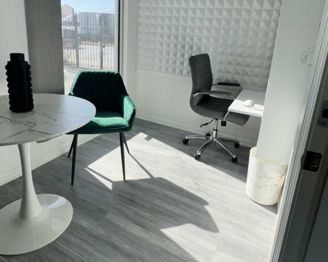 A small office space featuring a round table, a green chair, and a black office chair against a textured wall, with sunlight casting shadows on the gray flooring.