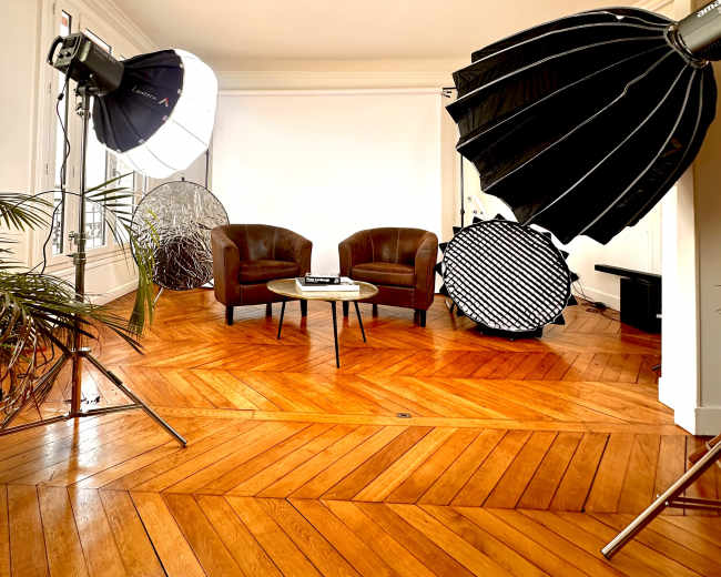 The image shows a photography studio with two brown chairs, a coffee table, and various lighting equipment set up on wooden flooring.