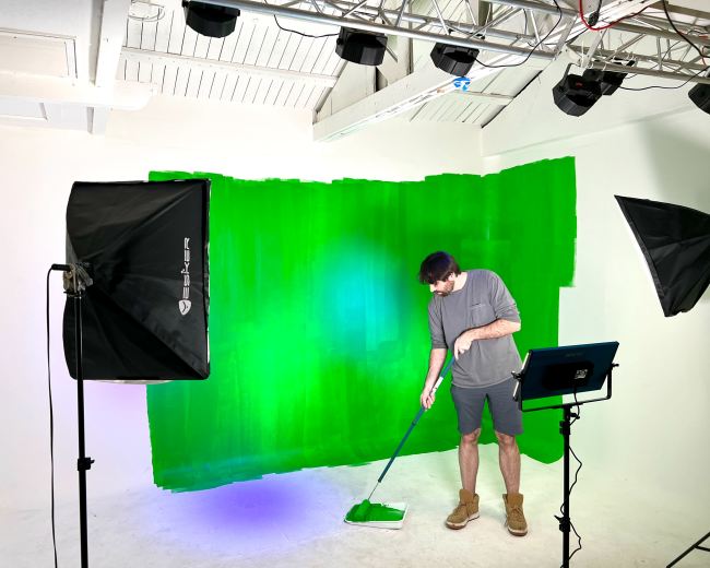 A person is painting a green backdrop in a photography studio while standing next to lighting equipment and a tablet.