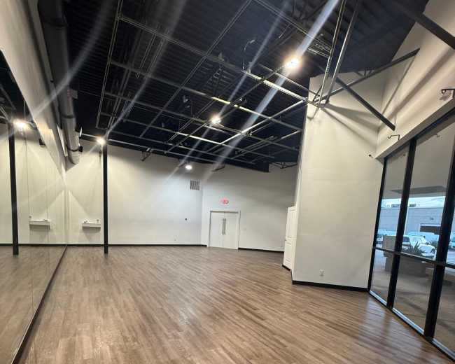 The image shows an empty room with wooden flooring, large mirrors on one wall, and bright overhead lighting.
