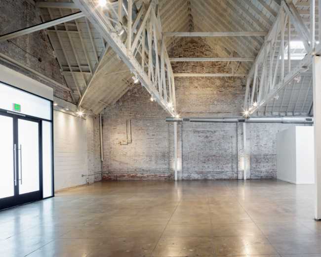 The image shows a spacious, empty interior of a modern building with high ceilings, exposed brick walls, and polished concrete flooring.