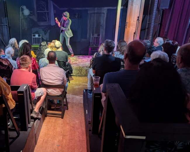 A performer in a suit plays guitar on stage while an audience watches from seated rows in a dimly lit venue.