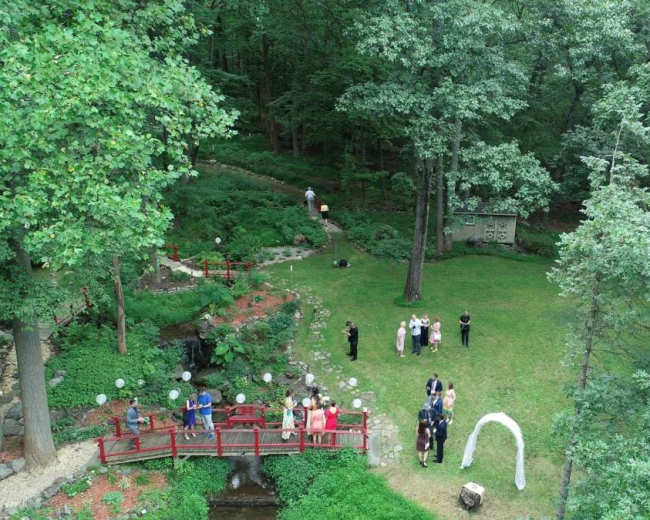 A wedding ceremony is taking place in a green garden, featuring a red bridge, a white arch, and guests gathered on the lawn.