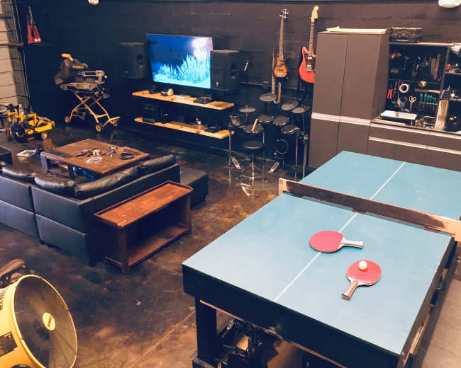 The image shows a multi-purpose room featuring two table tennis tables, a seating area with a black sofa and wooden coffee table, a large wall-mounted TV, musical instruments on display, and storage cabinets filled with various tools and equipment.