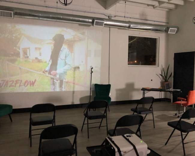 The scene shows a dimly lit room set up for a presentation, featuring chairs arranged in rows, a projector displaying an image of a person, and a small stage with a microphone.