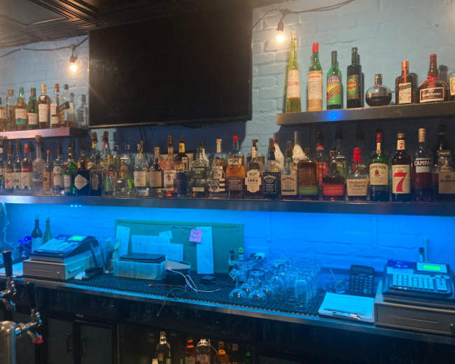 The image shows a bar counter with shelves lined with various bottles of liquor, and a blue ambient light illuminating the workspace below.