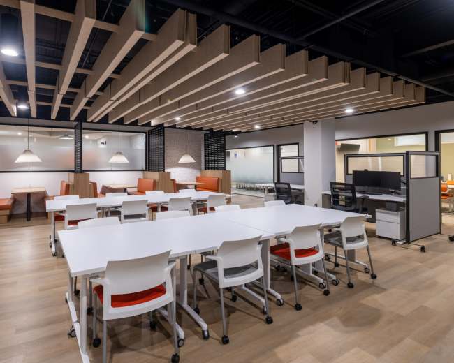 The image shows a modern office space with white tables and orange-accented seating areas, featuring a ceiling with wooden beams.