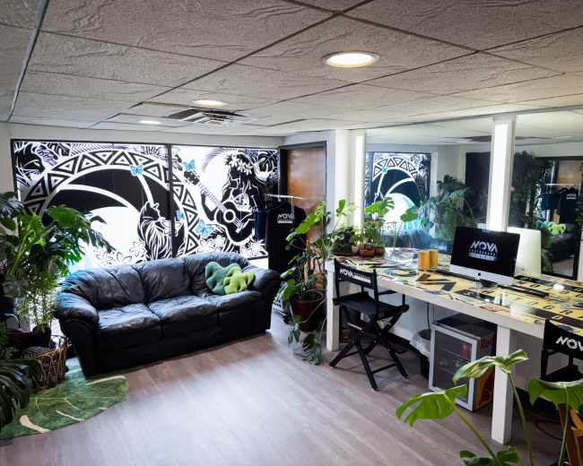 A cozy studio space featuring a black sofa, a desk with a computer, and plants, along with a wall decorated by a mural.
