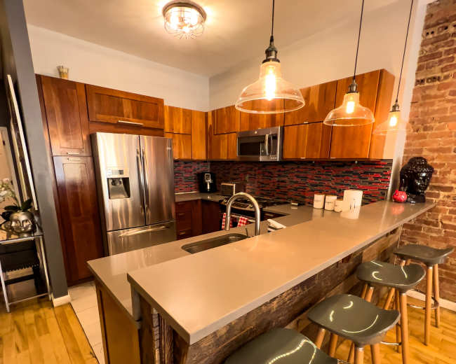 The image shows a modern kitchen with wooden cabinets, a gray countertop, an island with three stools, and exposed brick walls.