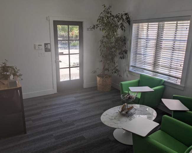 The image shows a bright, modern office space with green chairs, a marble coffee table, and a door leading to an outdoor area.