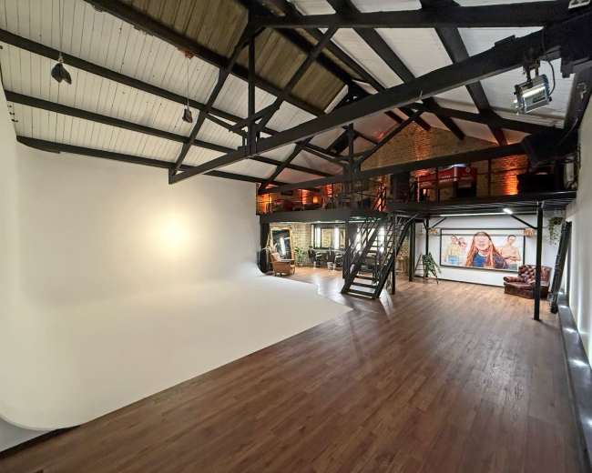 The image shows a spacious photography studio with a large white backdrop, wooden floors, and exposed beams, along with a staircase and an area featuring a large portrait on the wall.