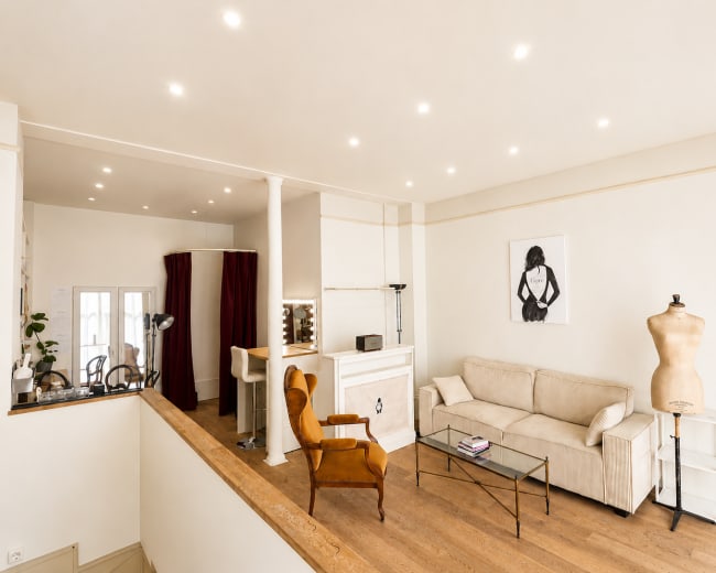 The image shows a bright, modern interior featuring a beige sofa, an orange chair, a glass coffee table, a mannequin, and a decorative poster on the wall.