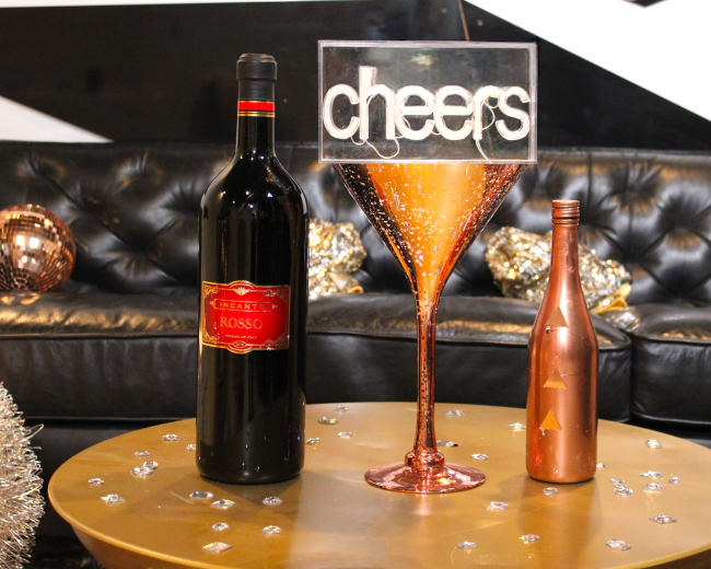 A large decorative martini glass filled with glimmering liquid stands on a gold table, surrounded by a bottle of red wine and a smaller bottle, against a backdrop of a black leather couch and metallic decor.
