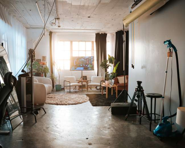 The image shows a spacious, well-lit studio with a mix of furniture, plants, and photography equipment arranged throughout the room.